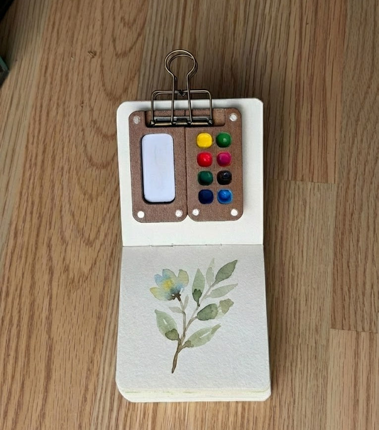 Miniature botanical watercolor painting in a small sketchbook with a portable wooden paint palette attached, resting on a wood desk.