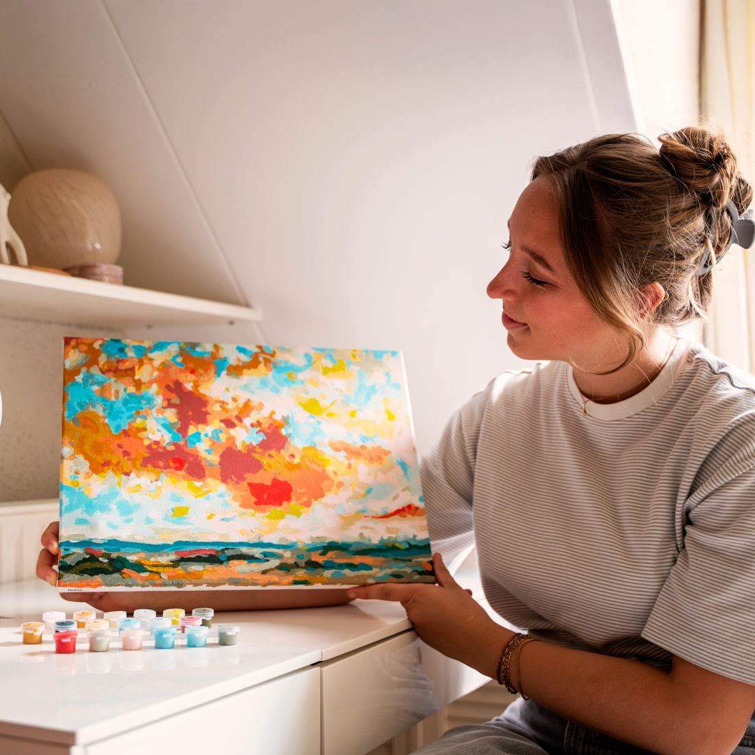 Woman smiling while holding a colorful abstract sunset landscape painting with small paint pots nearby