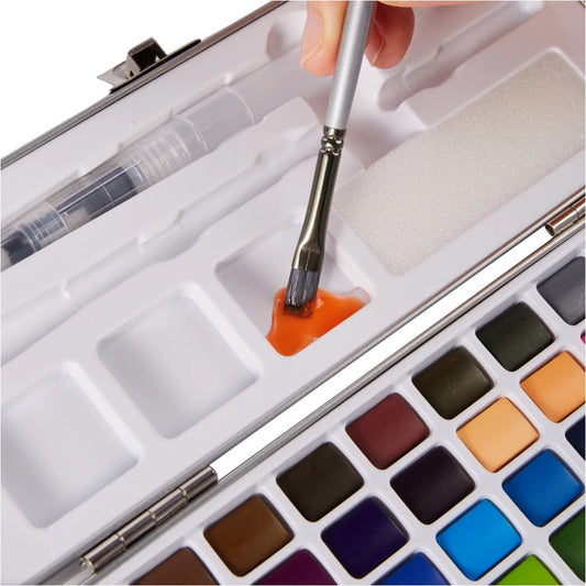 Brush mixing orange paint in a lid, mixing well beside solid watercolor pans in tin