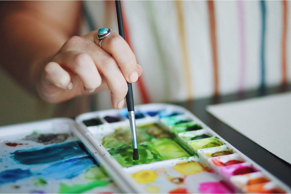 Closeup of a woman’s hand as she dips her paintbrush on paint in a white pallette