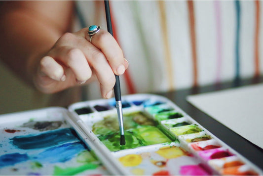 Closeup of a woman’s hand as she dips her paintbrush on paint in a white pallette