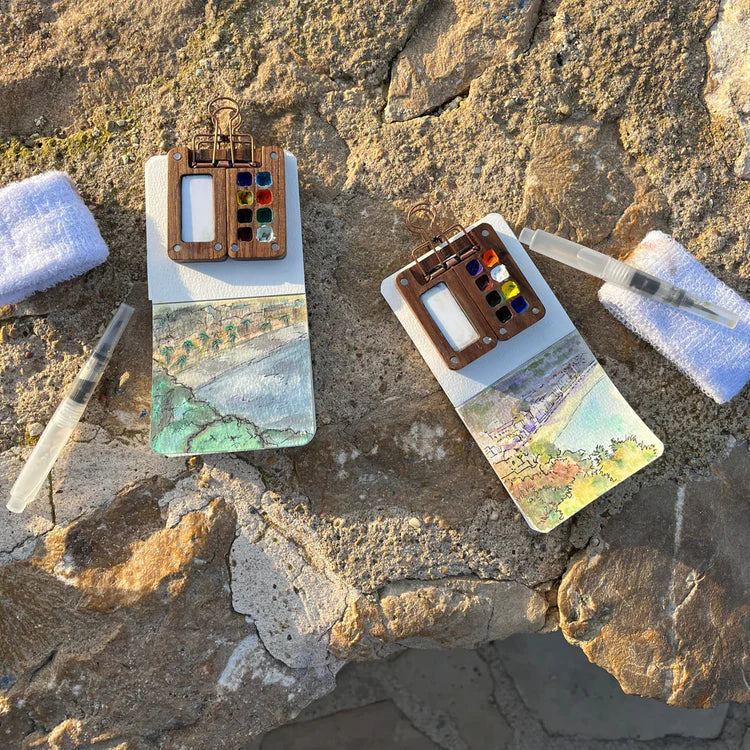 Two compact portable watercolor painting kits with landscape sketches displayed on sunlit rocks outdoors.