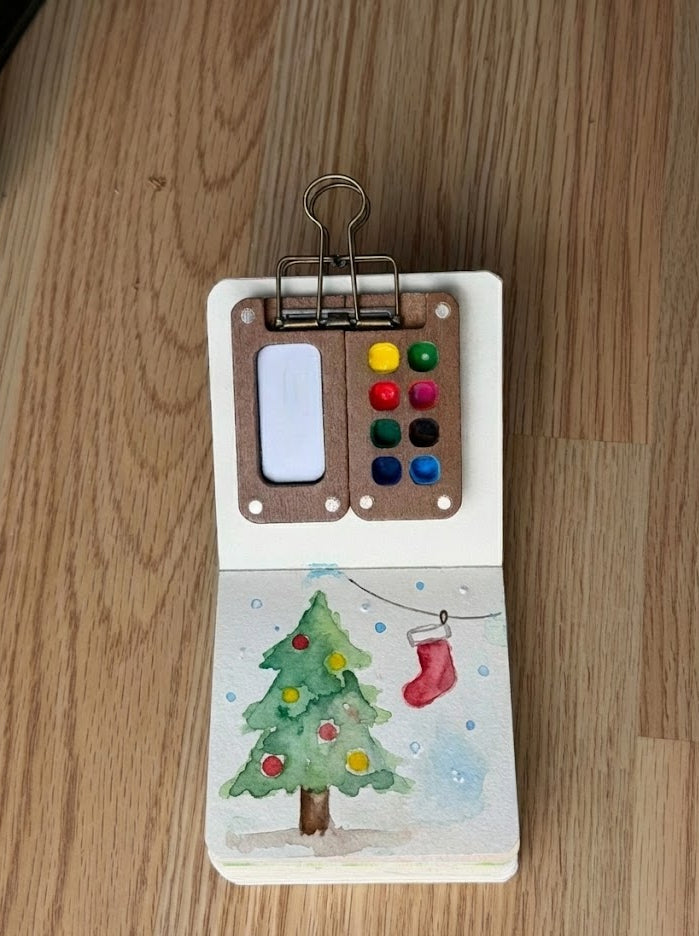 Watercolor painting with a Christmas tree and colorful palette on a wooden surface