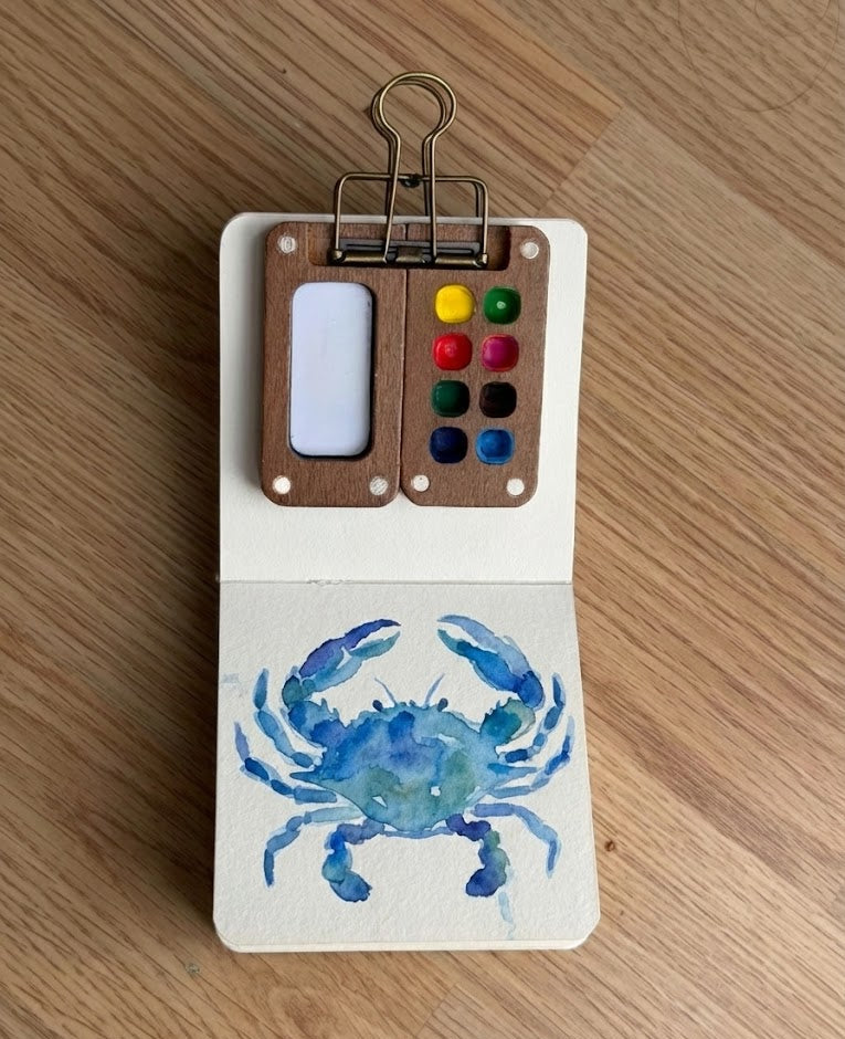 Art kit with watercolor paint set and blue crab illustration on a wooden surface