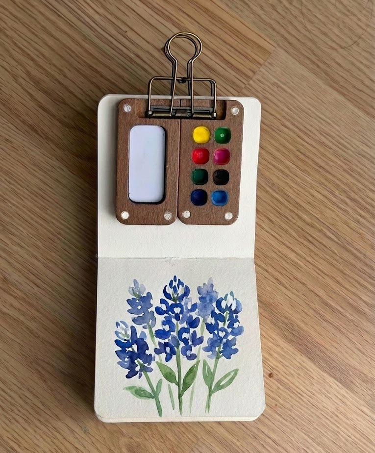 Small wooden art palette with paint swatches and a clip, featuring bluebonnet watercolor painting on a wooden surface.