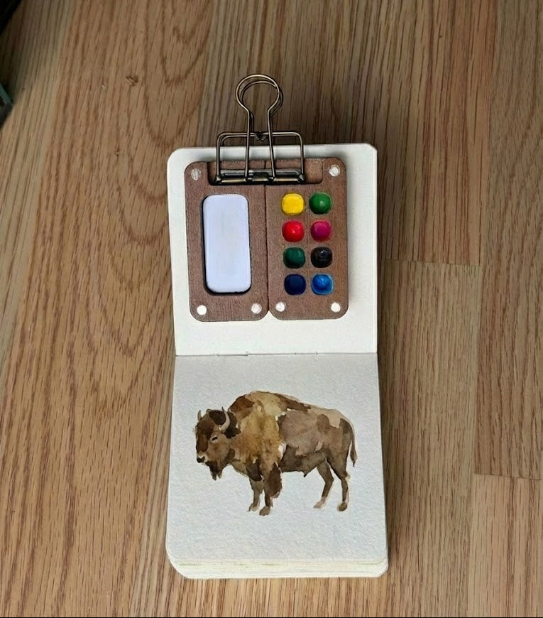 A miniature watercolor painting of a brown bison in a small sketchbook. A tiny wooden palette with eight colorful paint wells is clipped to the top page, resting on a light wood floor.
