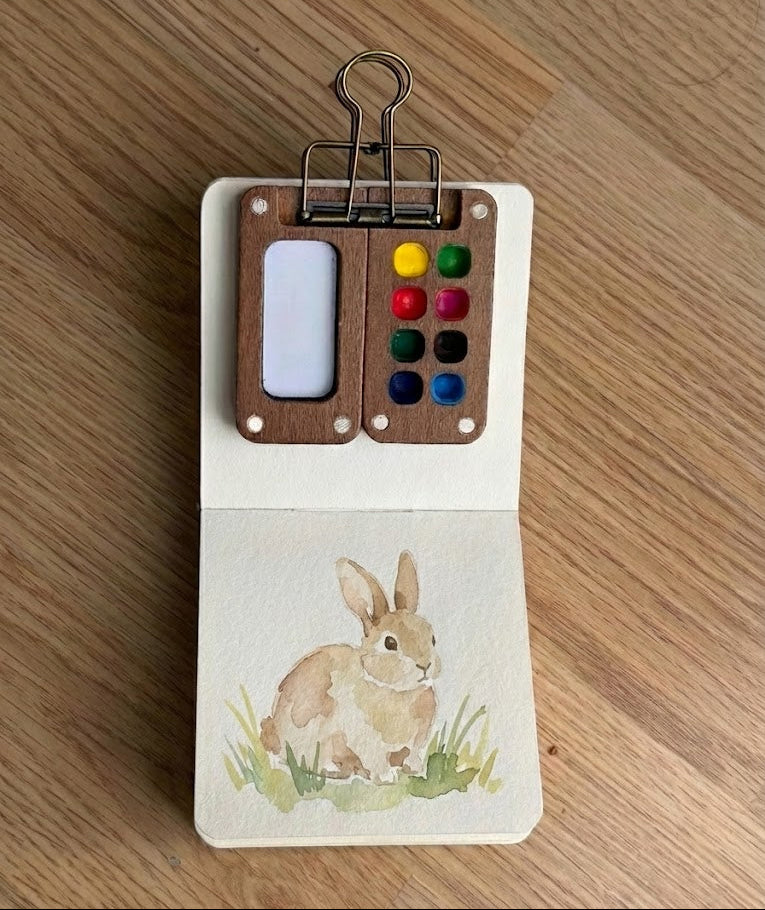 Small wooden art set with watercolor paints and a bunny watercolor painting on a wooden surface