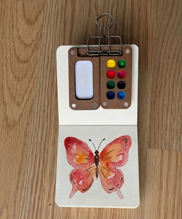 Painting of a butterfly on a small art board with watercolor paints on a wooden surface