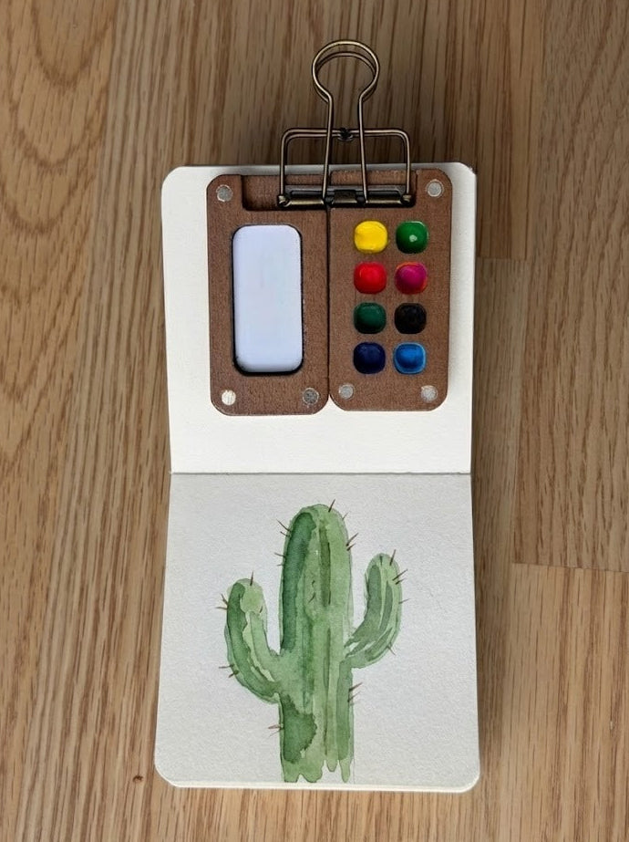 Small sketchbook open on a wooden table displaying a green saguaro cactus watercolor painting