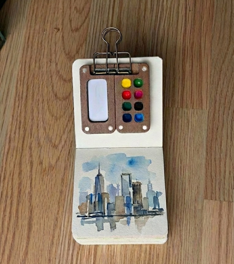 Small open book with watercolor cityscape and colorful buttons on a wooden surface