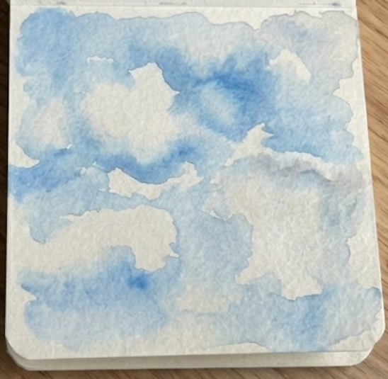 Cerulean Blue watercolor paint dropped into the wet sky wash on a sketchbook page, defining the initial white cloud shapes.