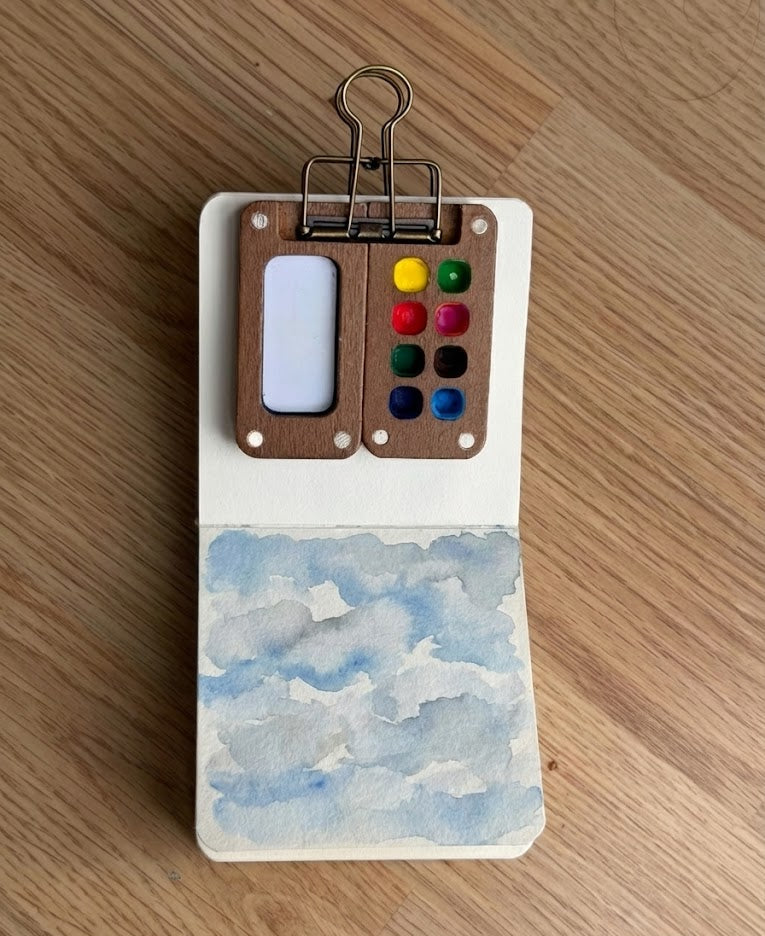 Hand-painted blue and grey watercolor clouds in a pocket sketchbook on a wooden desk.