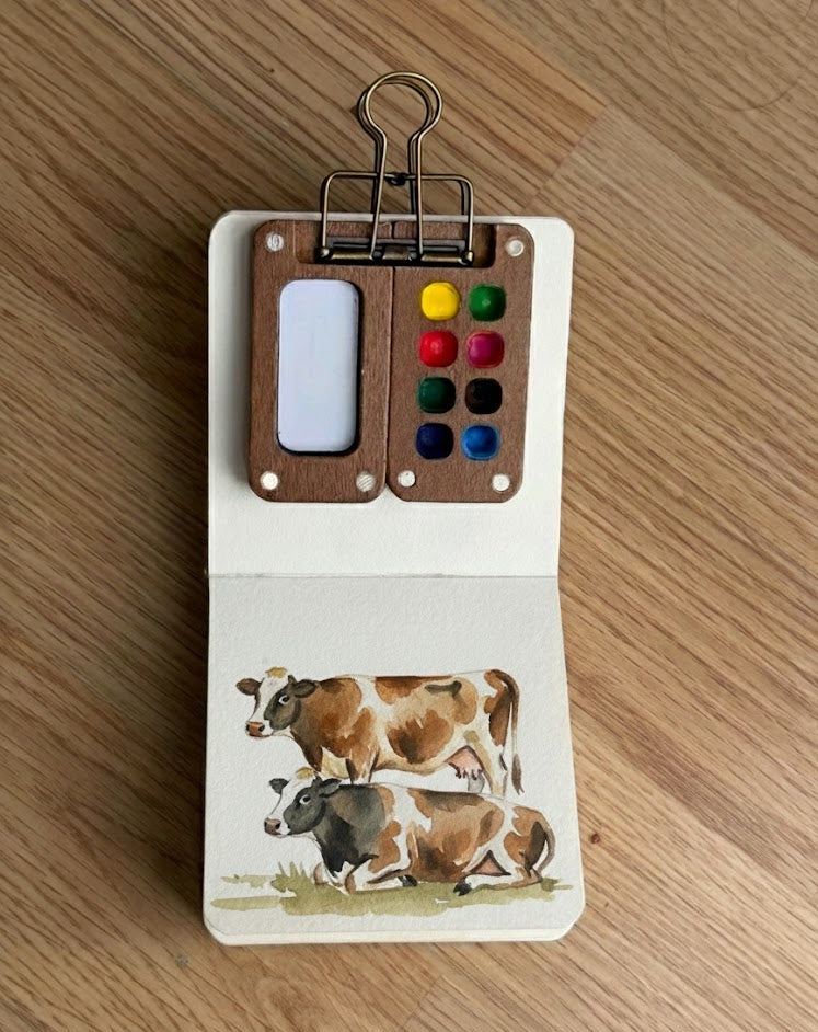 Mini wooden travel watercolor palette with six paint wells clipped to a small sketchbook showing a cow illustration.