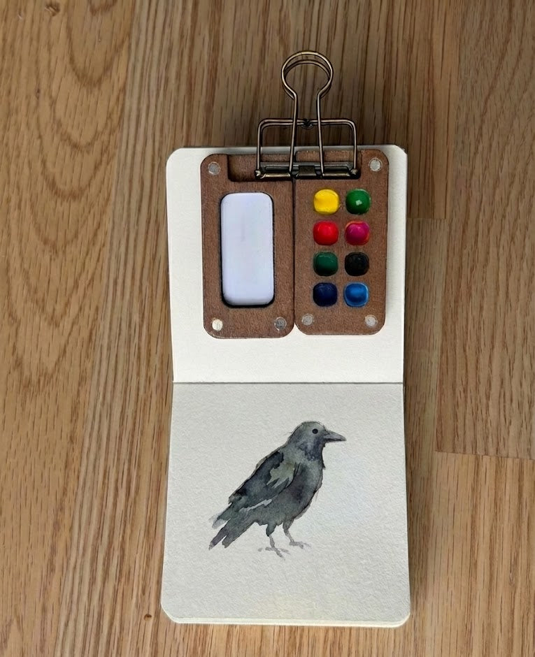 Watercolor paint palette and illustration of a crow on wooden surface