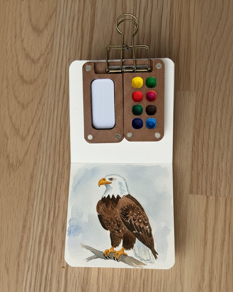 An open sketchbook featuring a watercolor painting of a bald eagle, with a compact wooden paint palette clipped to the top page.
