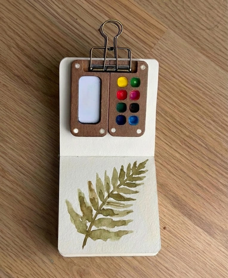 A watercolor painting of a green fern inside a small open sketchbook with a tiny wooden paint palette clipped to the top.