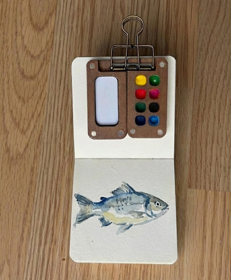 Small watercolor painting of a fish on a wooden surface with a color palette.