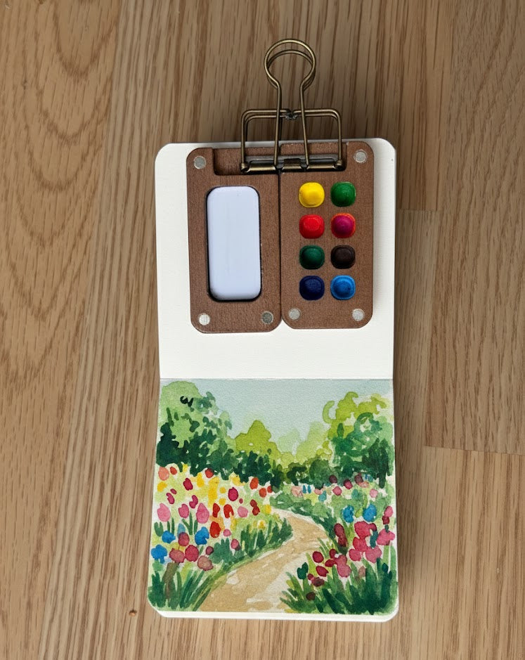 Small paint palette with colorful buttons and a painted scene of a garden on a wooden surface