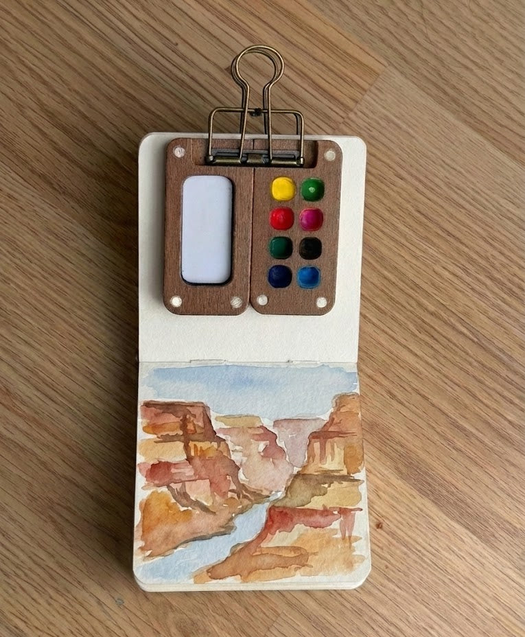 A small, impressionistic watercolor painting of the Grand Canyon inside a sketchbook, featuring layered rock formations in earthy tones of terracotta, ochre, and sienna under a pale blue sky.