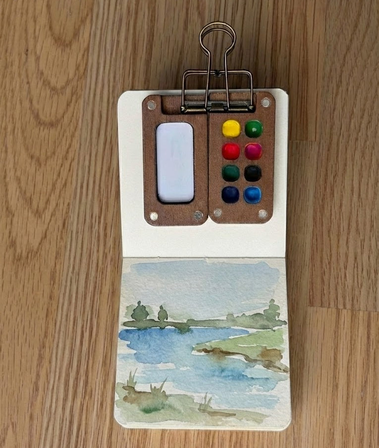 Small watercolor painting of a lake on a wooden surface with a miniature paint palette.