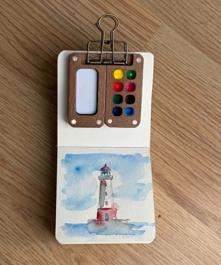 Small watercolor painting of a lighthouse on a wooden surface with a paint palette clip.