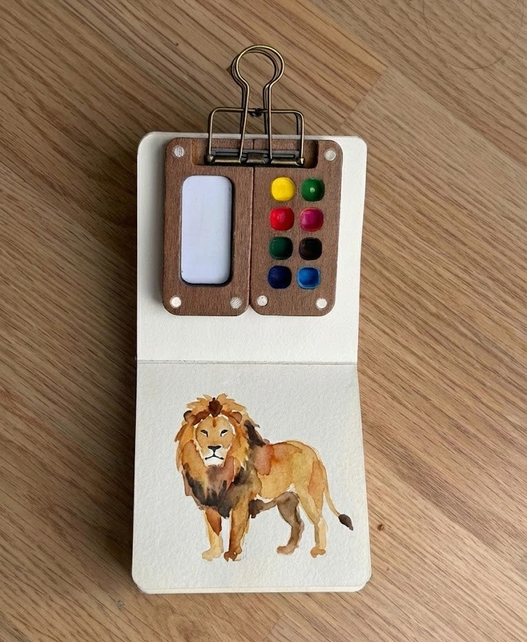Small book with a lion watercolor painting and a clip on a wooden surface