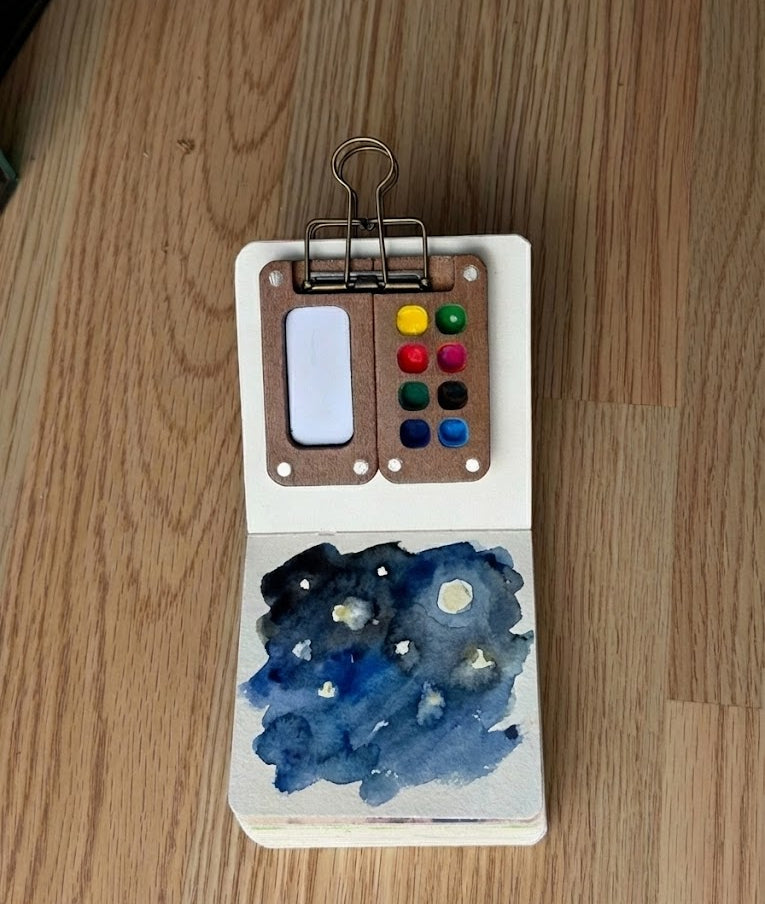 Watercolor painting of night sky with watercolor pages and a clip on wooden surface