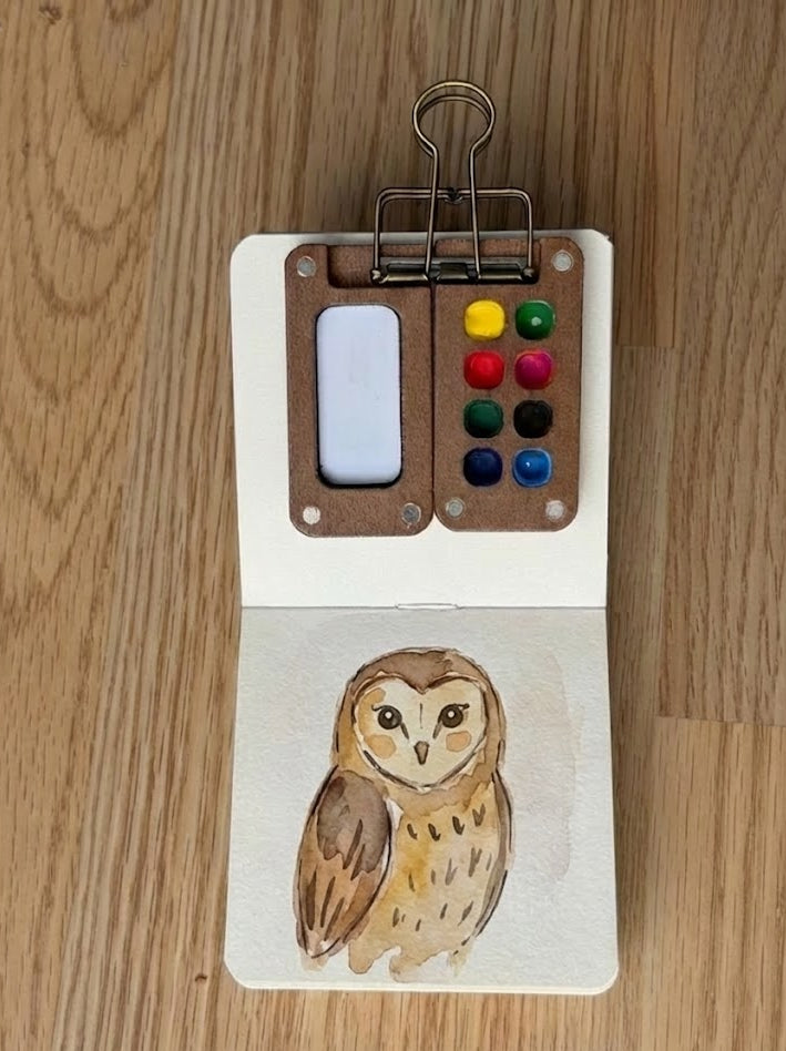 Small watercolor painting of an owl on a wooden surface with a miniature paint palette.