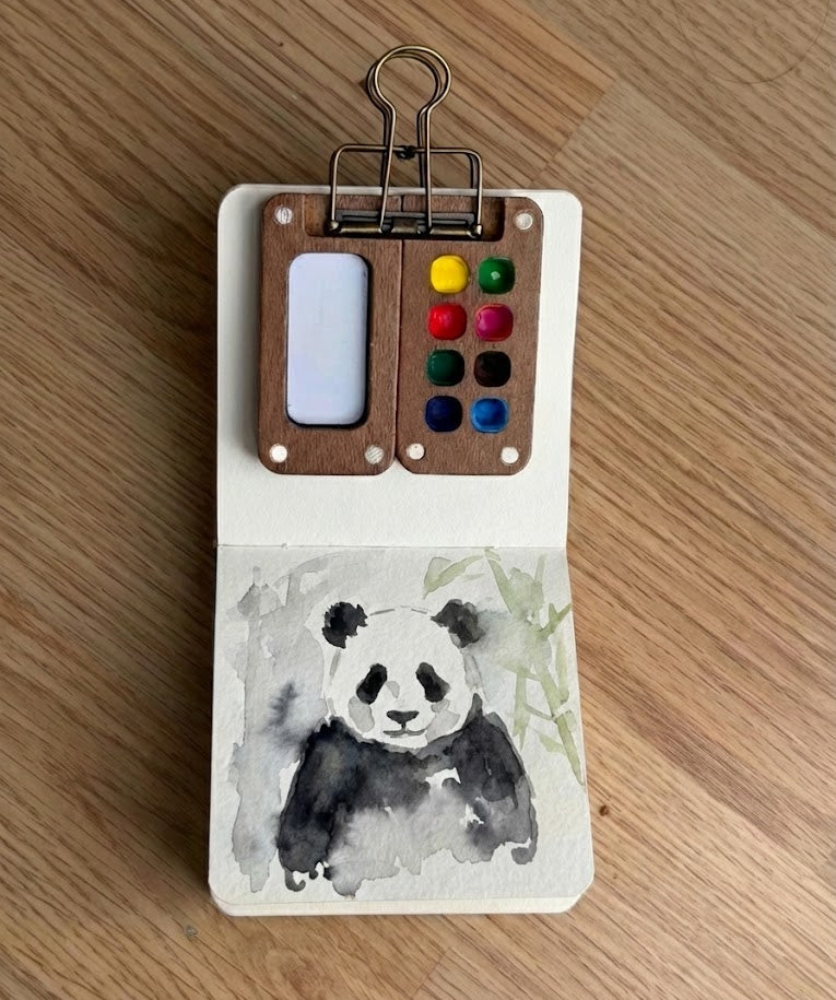 Small art kit with watercolor paints and a panda illustration on a wooden surface