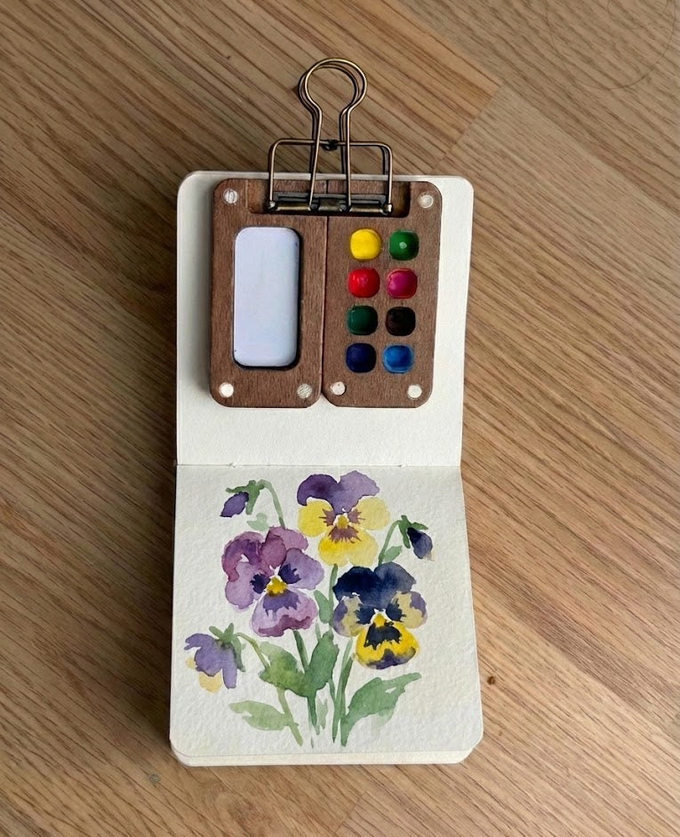 Watercolor painting of pansies in a mini sketchbook with a tiny wooden paint palette clipped to it.