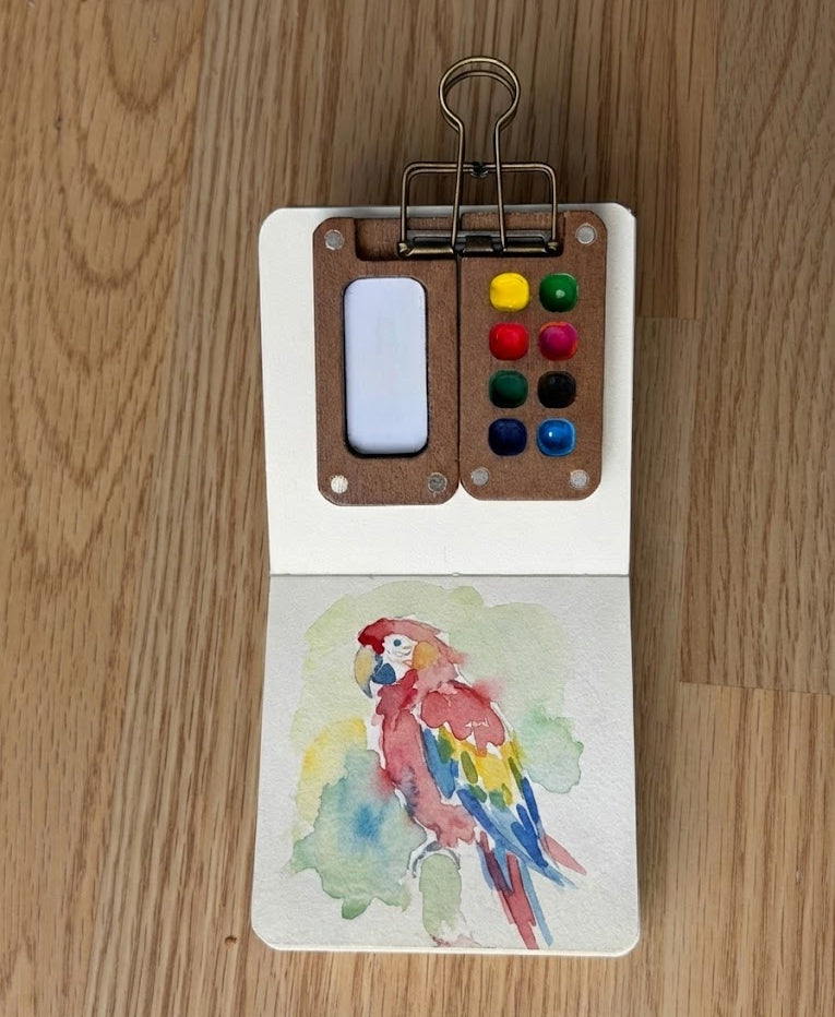 Small watercolor book with a parrot watercolor painting and color swatches on a wooden surface