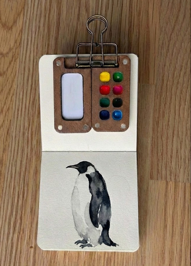 Small book with a penguin watercolor painting and color swatches on a wooden surface