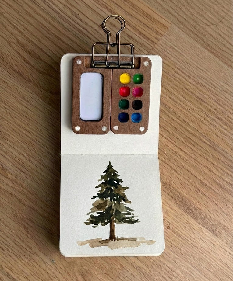 Small watercolor book with a tree painting and paint palette on a wooden surface