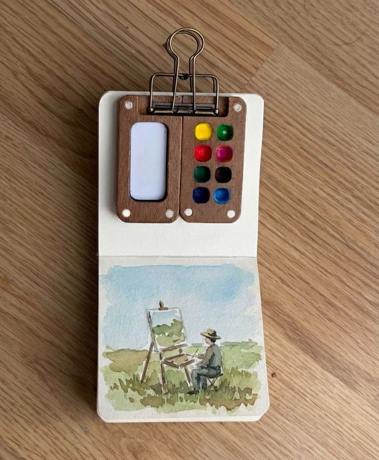 Small art kit with watercolor paints and a painting of plein air on a wooden surface.