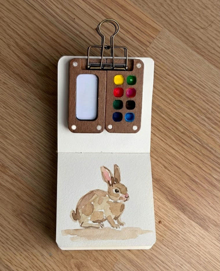 Small paint palette with watercolor paints and a painting of a rabbit on a wooden surface