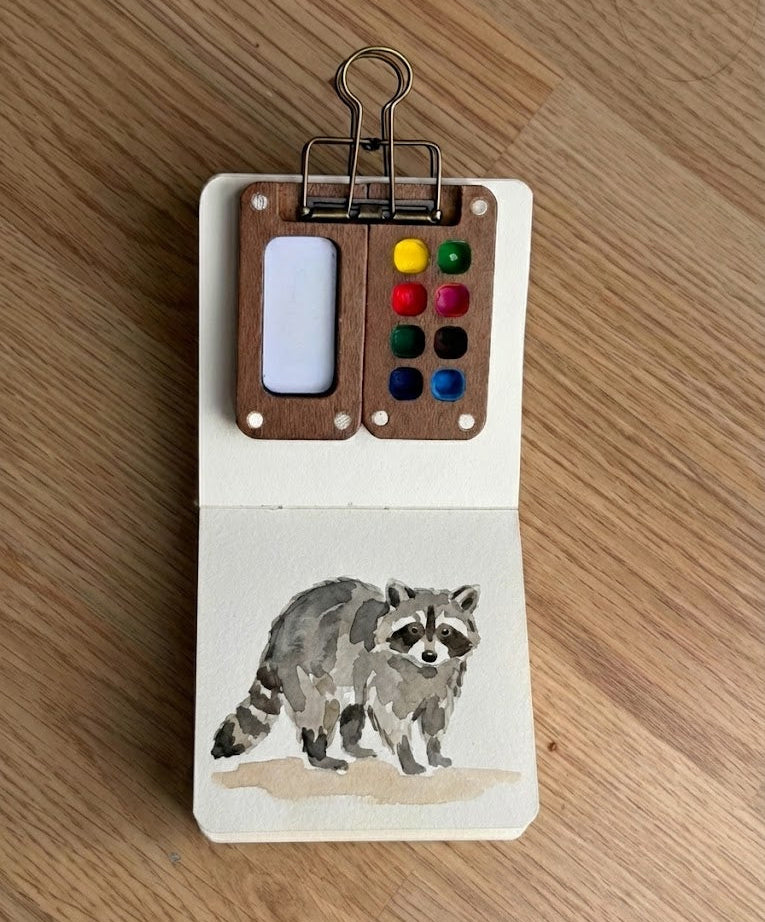 A raccoon illustration and a paint palette on a wooden surface