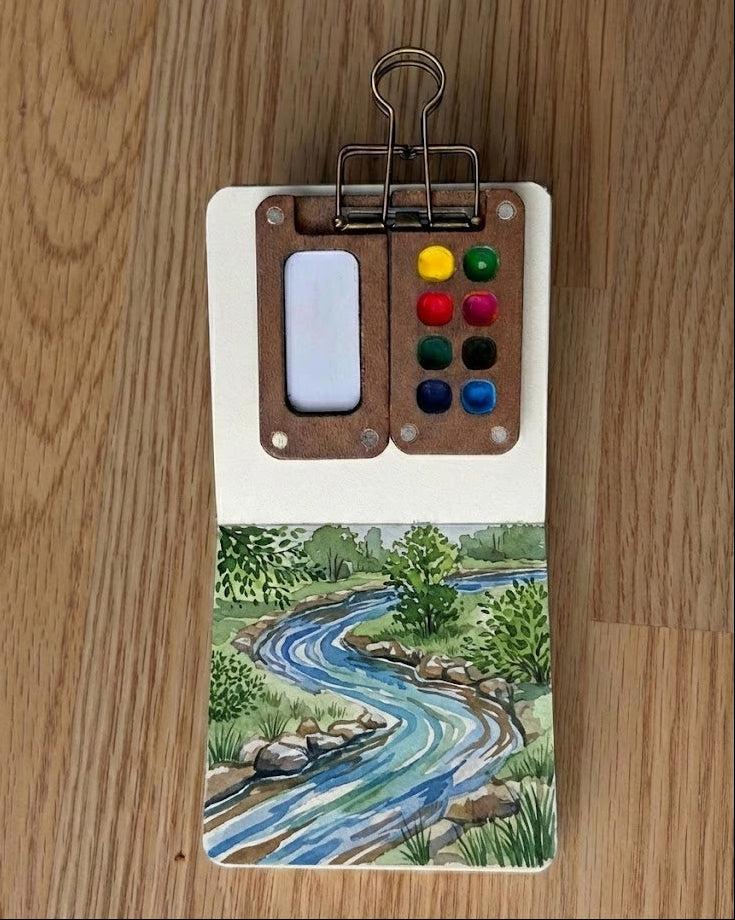 Small watercolor painting of a river scene with a color palette on a wooden background