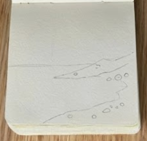Initial pencil sketch of a horizon line and sloping beach in a small sketchbook, representing step one of a step-by-step seascape watercolor painting.