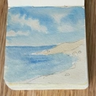 Adding a wash of blue sky and leaving dry patches for clouds in a sketchbook, showing step two of a seascape watercolor painting.