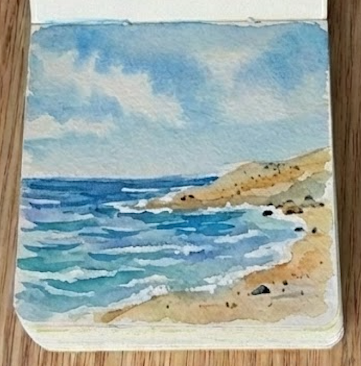 The completed seascape watercolor painting in a mini sketchbook with an attached wooden paint palette, featuring a finished sandy beach, ocean waves, and dark rocks.