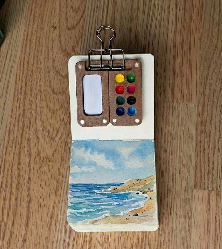 A small watercolor painting in a sketchbook depicting a bright seascape. Blue and turquoise waves crash against a curved, sandy shore under a light blue sky with fluffy white clouds.