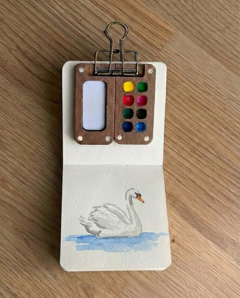 A top-down view of a small open sketchbook featuring a delicate watercolor painting of a white swan, with a miniature wooden paint palette clipped to the top page.