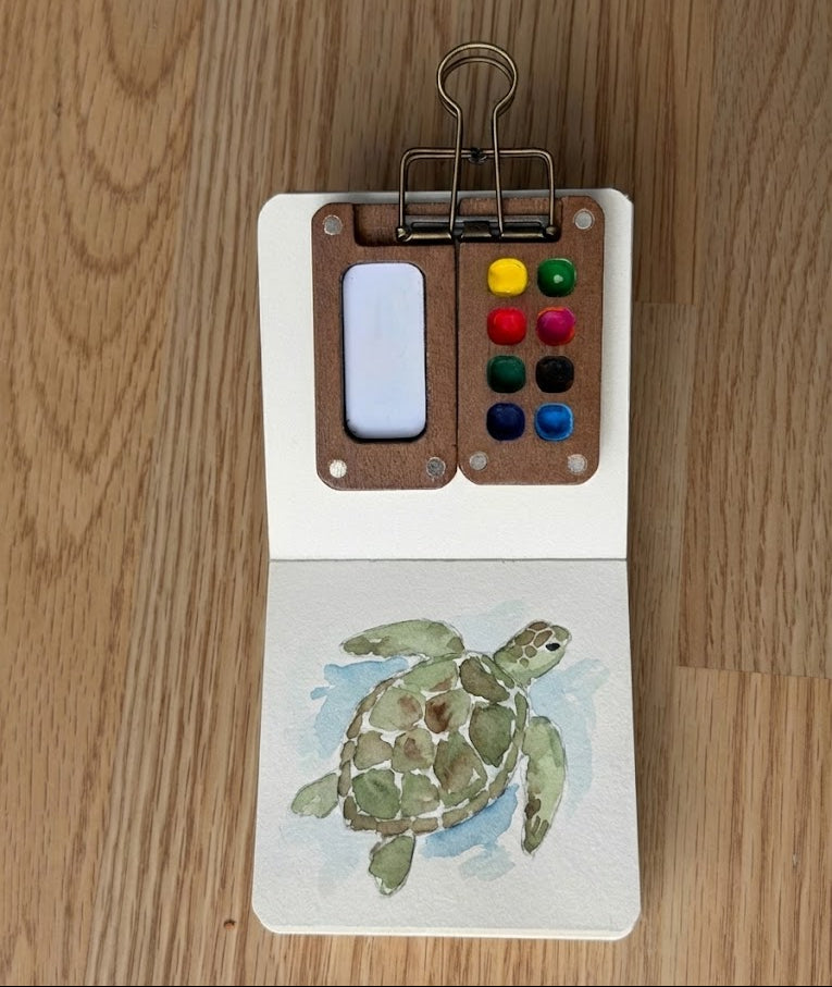 Small watercolor painting of a turtle on a wooden surface with a paint palette and brush.