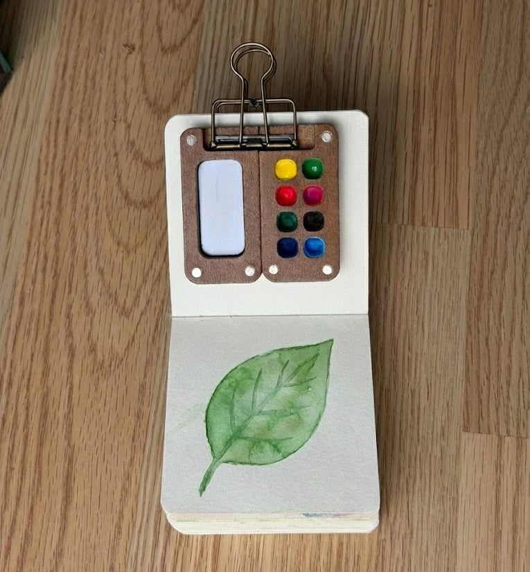 Miniature watercolor painting of a green leaf in a small open sketchbook, featuring a tiny wooden travel watercolor palette attached by a binder clip.